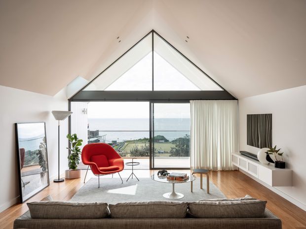 A bright, refined living room with a dramatic vaulted ceiling and triangular clerestory glazing above a large black-framed window wall. The room has warm timber flooring, a neutral sectional sofa, a vivid red lounge chair, a white built-in media console, soft curtains, and views toward the ocean and landscaped grounds. A large mirror leans against the wall, reflecting the coastal outlook.