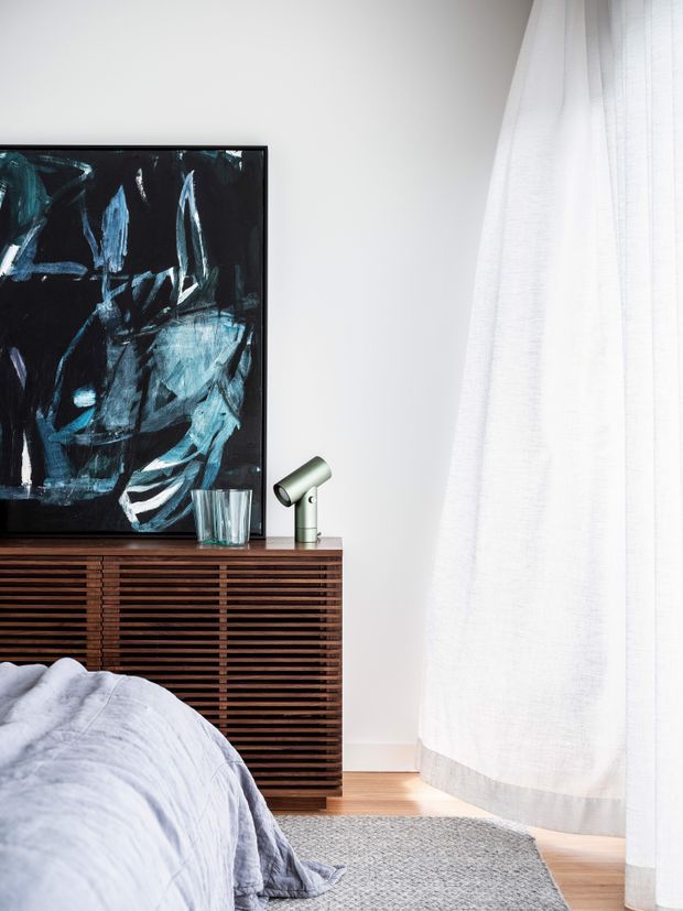 A softly lit contemporary bedroom corner featuring a low dark timber slatted sideboard, abstract black-and-blue artwork, a small metallic table lamp, and a bed with pale grey-blue bedding. Sheer white curtains cover a tall window, with light timber flooring and a textured grey rug creating a calm, refined atmosphere.