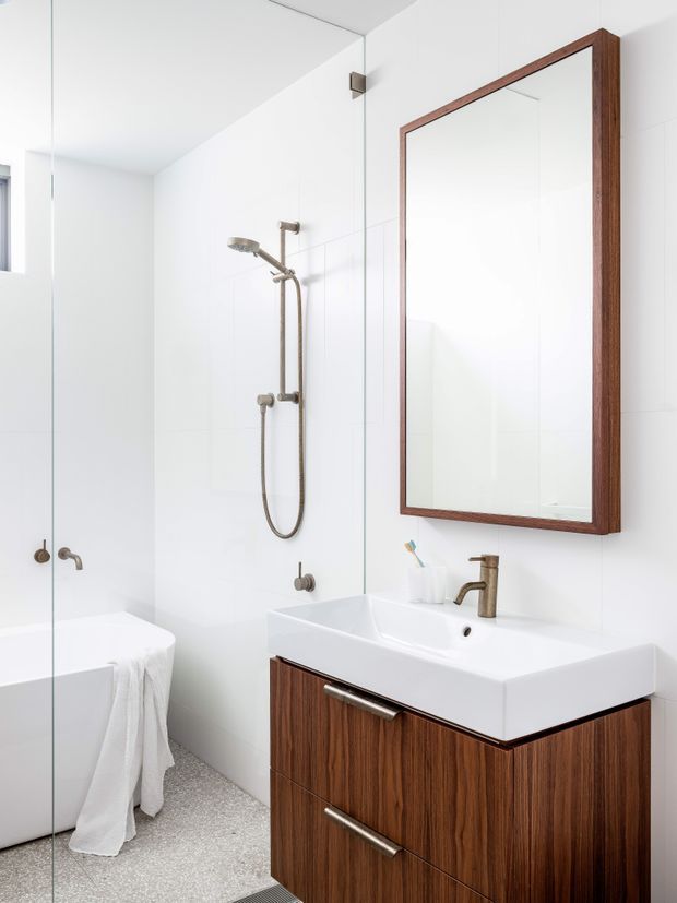 Bright, clean contemporary bathroom with a white tiled shower enclosure, freestanding bathtub partially visible at left, wall-mounted white basin, and dark timber vanity cabinetry. A large rectangular timber-framed mirror, brushed metal shower fittings, and terrazzo-like grey speckled flooring complete the minimalist fit-out.