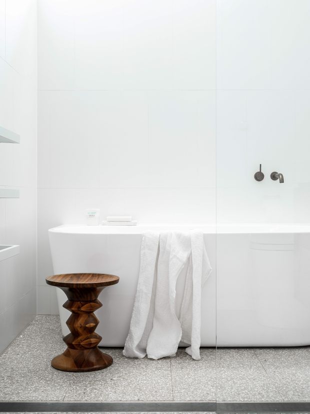 Bright, minimalist white bathroom featuring a freestanding bathtub, large white wall tiles, terrazzo-style grey flooring, and dark metal wall-mounted bath controls. A sculptural dark timber stool and white towels add warmth and texture.