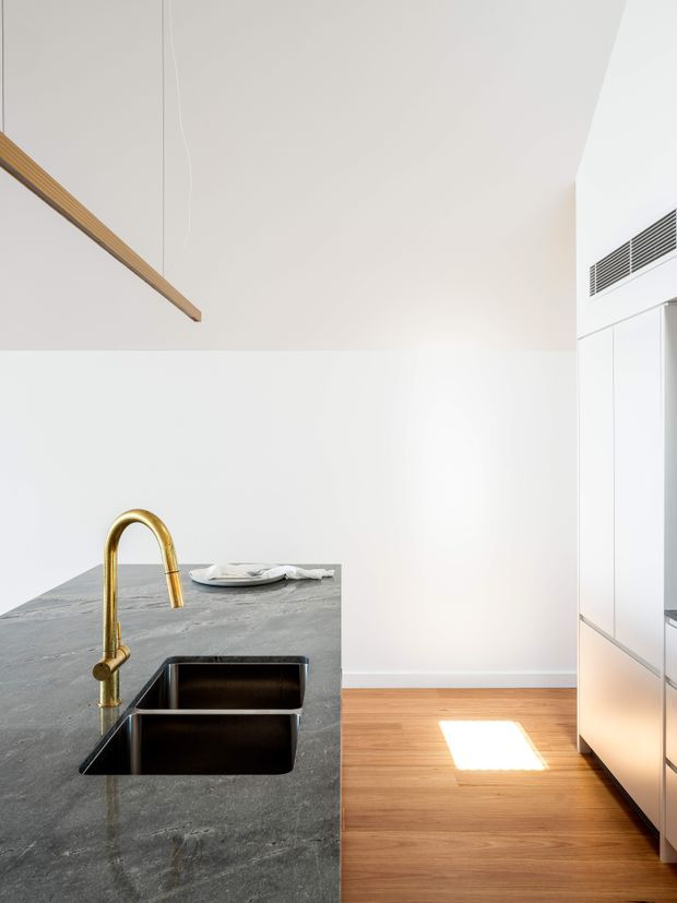 Bright minimalist kitchen interior with a large dark grey stone island, integrated double stainless-steel sink, and a prominent brushed gold gooseneck tap. White walls and ceiling, pale timber flooring, white full-height cabinetry, and linear timber pendant lighting create a clean contemporary composition. A high-level air-conditioning grille is visible on the right wall.