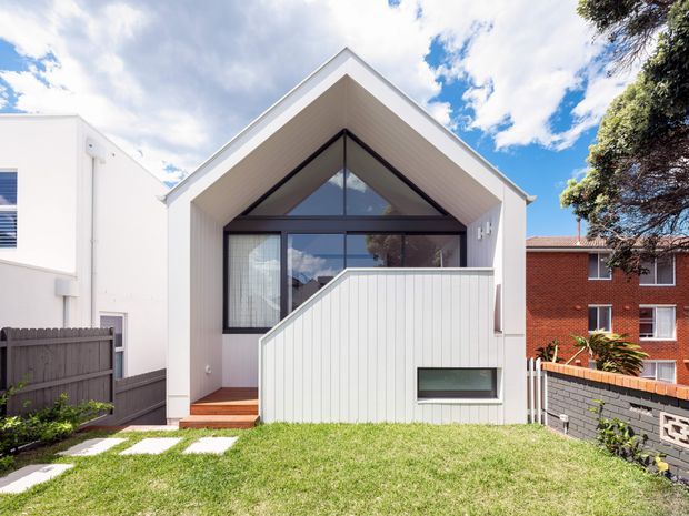 Front exterior view of a contemporary white-clad house extension or compact dwelling with a prominent steep gabled roof and triangular clerestory glazing. The facade combines vertical light-coloured cladding, dark-framed windows, a raised timber entry deck, and a solid privacy wall with a horizontal window. Neighbouring homes, boundary fencing, lawn, and stepping-stone landscaping are visible.