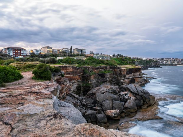 Coastal cliffside landscape with layered sandstone rock platforms and a rugged rocky inlet in the foreground. Low-rise residential buildings line the elevated shoreline in the background, with ocean surf along the right side beneath an overcast sky.