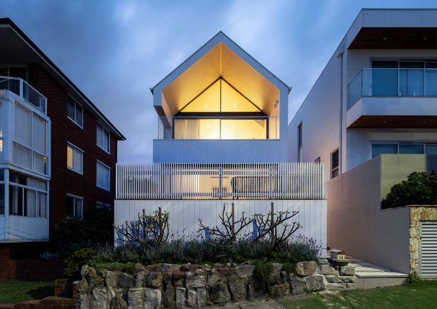 Exterior dusk view of a narrow contemporary multi-storey residence between neighbouring homes. The central house has a prominent front-facing gabled roof with a warm illuminated timber-lined triangular soffit, large upper-level glazing, a white rendered facade, and a slatted metal privacy screen enclosing the lower level. Adjacent modern balconies and landscaped stone retaining walls are also visible.