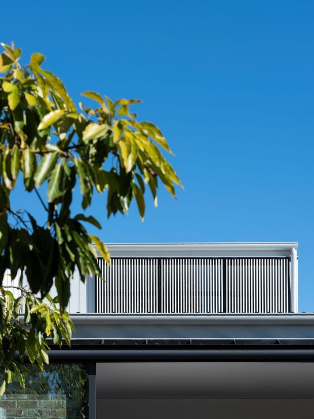 Partial exterior view of a contemporary multi-level residence under a clear blue sky. The facade features a flat roofline, crisp white horizontal trim, dark charcoal surfaces, and a prominent upper-level screen of closely spaced vertical slats. Mature leafy planting partially obscures the lower left of the building.