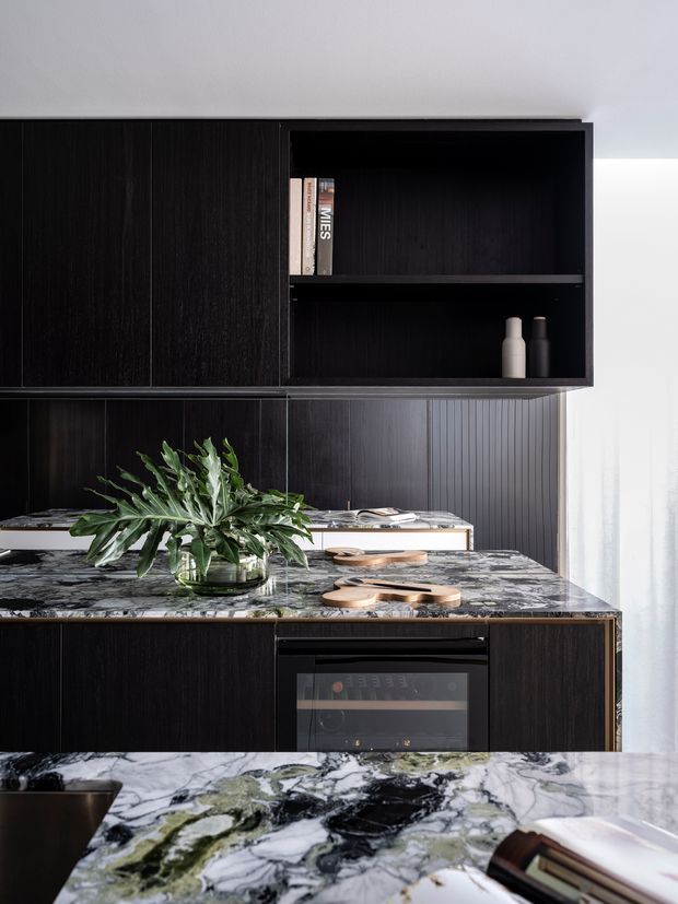 A sophisticated contemporary kitchen dominated by dark vertical-grain timber cabinetry and a dramatic open shelving unit. Veined natural stone surfaces with black, white and green marbling form the island and counters, with an integrated wine cooler or appliance below. Decorative bottles, books, timber serving boards and a leafy plant add restrained styling. Bright daylight enters from the right through sheer curtains, contrasting with the dark cabinetry.
