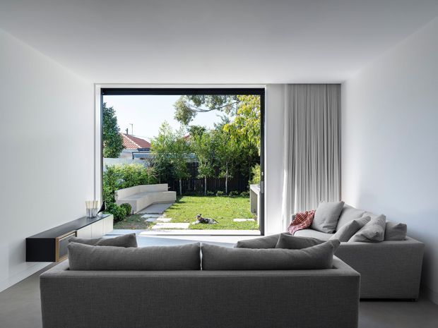 Bright, minimalist living room viewed from behind a large grey sectional sofa. The room has white walls and ceiling, a pale smooth floor, a low dark built-in media cabinet, and full-height light grey curtains beside a large black-framed opening to a landscaped backyard. A second grey sofa and simple side table complete the restrained contemporary interior.