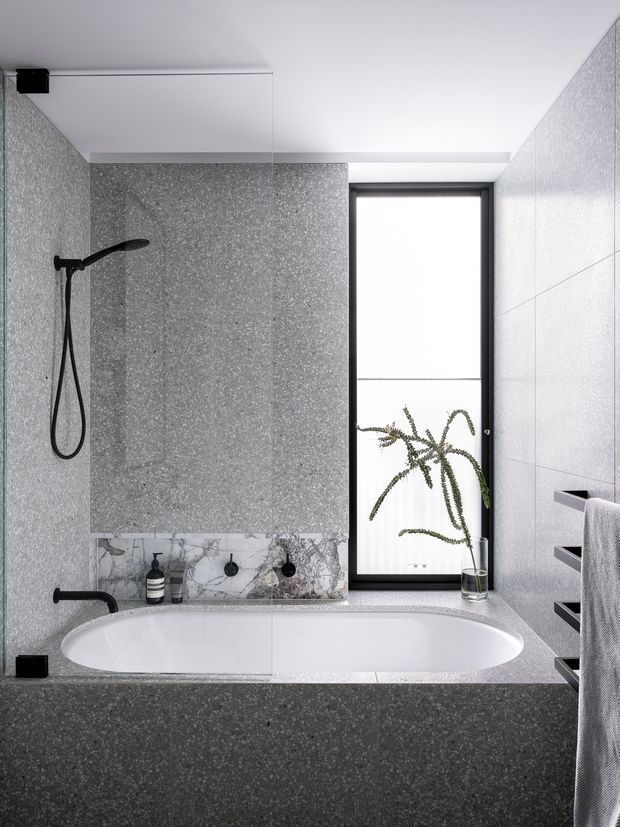 A contemporary bathroom featuring a built-in white bathtub set into a grey terrazzo-like surround, large grey textured wall tiles, black shower and tapware, a narrow black-framed window, and recessed black shelving with a towel. A small plant and toiletries sit on the bath ledge. The space has a clean, monochromatic, refined look with strong textural contrast.