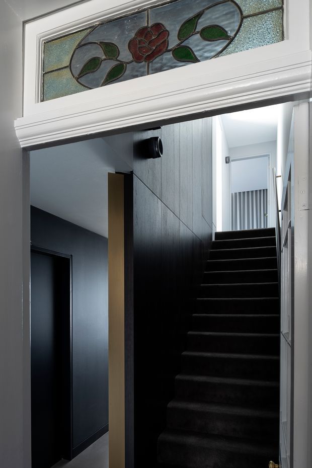 Interior view through a traditional doorway into a dark, narrow stairway. The staircase is fully carpeted in deep charcoal or black, with dark vertical wall panelling and a contrasting pale timber or painted trim element. A decorative stained-glass transom window with floral and leaf motifs sits above the doorway, while a brighter upper landing is visible beyond the stairs.