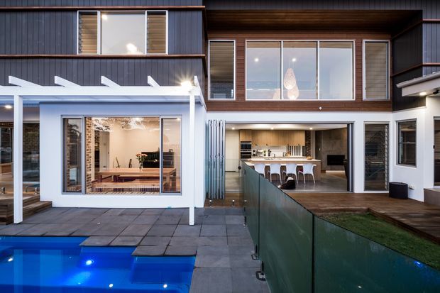 Contemporary two-storey residential facade viewed from the pool courtyard, with dark vertical cladding, timber-lined upper walls, large windows, a covered outdoor sitting area, and an open-plan kitchen visible through the glazing. A tiled pool terrace and glass pool fence occupy the foreground.