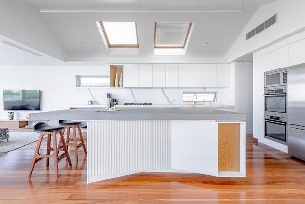Bright, contemporary open-plan kitchen with a large white island, integrated appliances, white flat-panel cabinetry, marble-look splashback, timber accent joinery and a ribbed island façade. Two skylights provide abundant natural light. Timber flooring continues into the adjacent living area, which includes a television and seating.