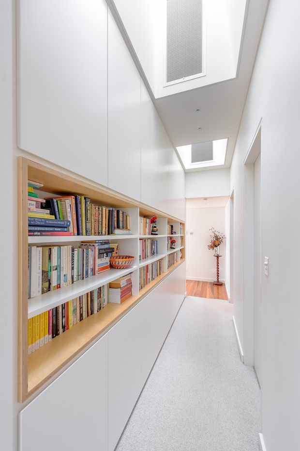 A bright, narrow interior hallway lined with extensive built-in white cabinetry and open timber-edged bookshelves filled with books. The corridor has a light carpeted floor, white walls and ceiling, recessed downlights, and multiple rectangular skylights or roof lights. A timber-floored room is visible at the far end, with a decorative vase and dried branches. The overall design is clean, minimalist, and highly functional.