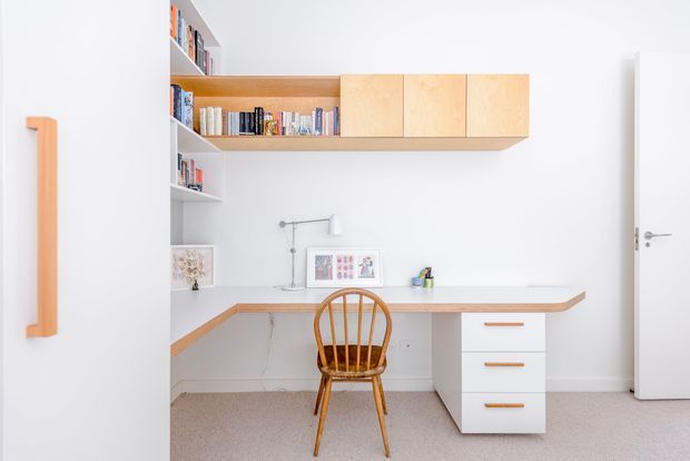 Bright, minimalist home office with a built-in L-shaped timber desk, white drawer pedestal, open book shelving, and pale timber overhead cabinetry. A spindle-back wooden chair sits at the workspace, with white walls, a light carpeted floor, and simple wall-mounted task lighting.