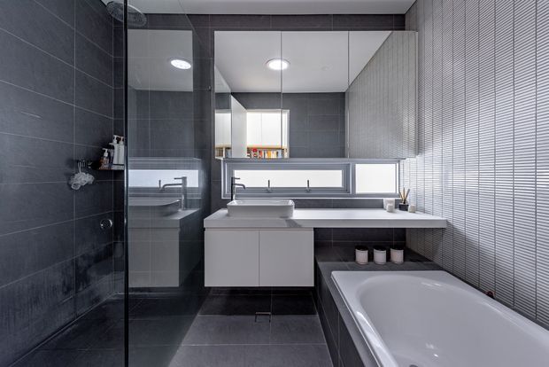 Contemporary bathroom featuring dark large-format tiled walls and floor, a frameless glass shower enclosure, floating white vanity with integrated basin, wall-mounted mirror cabinetry, a freestanding-style bathtub, and horizontal windows with venetian blinds. The palette is monochromatic grey, black, and white with clean-lined fixtures and strong natural light.