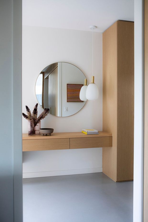 A minimalist entryway or hallway vignette with a floating light-wood console, integrated tall timber cabinetry, a large round mirror, and a sculptural white pendant light with brass detail. The room has smooth white walls, a pale grey seamless floor, and restrained decorative objects including dried foliage, a bowl, and books. The mirror reflects an adjacent bright space and artwork.
