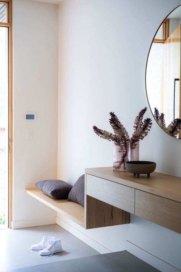 Bright minimalist entryway with a built-in light-wood bench and grey cushions beside a floating light-wood console. A large round black-framed mirror, ribbed glass vases with dried foliage, wall controls, timber-framed glazing, and pale flooring create a clean, contemporary interior.