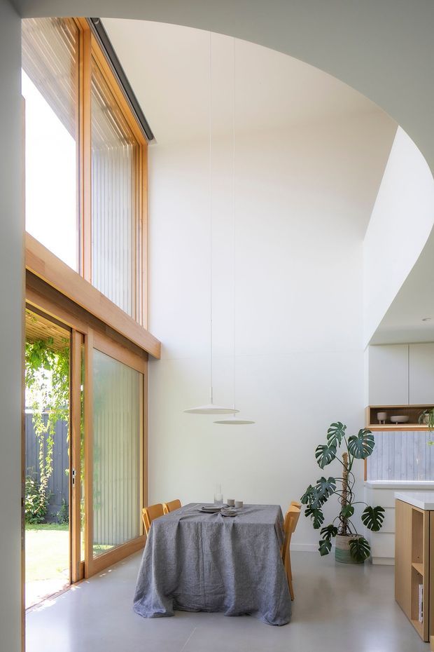 Bright, minimalist dining area within an open-plan home, featuring a round dining table dressed in grey linen, timber-framed floor-to-ceiling glazing and sliding doors, white walls, a dramatic curved high ceiling, sculptural pendant lights, and a leafy indoor plant. A compact kitchen with white cabinetry and timber joinery is visible to the right. The space has a calm, airy, contemporary character with strong indoor-outdoor connection.