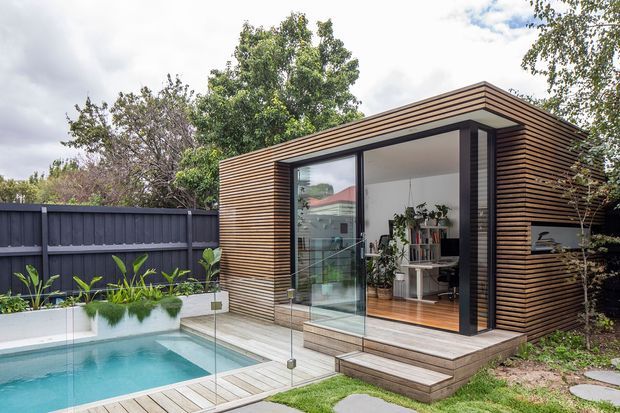 A compact contemporary garden pavilion beside a small swimming pool. The pavilion has horizontal timber-look cladding, a projecting flat roof, extensive floor-to-ceiling glazing with dark frames, and timber steps leading into a bright plant-filled interior. Glass pool fencing, raised garden planters, lawn, and a dark boundary fence surround the structure.