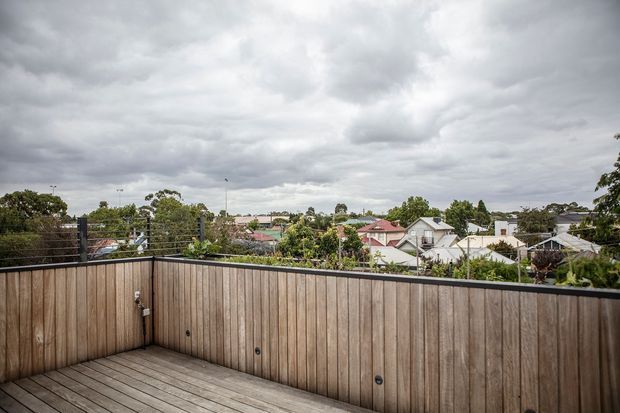 A rooftop or elevated timber deck overlooking a suburban residential neighbourhood. The deck is enclosed by a tall vertical timber privacy balustrade with dark metal posts and wire railing above, while the surrounding homes and trees sit beneath an overcast sky.