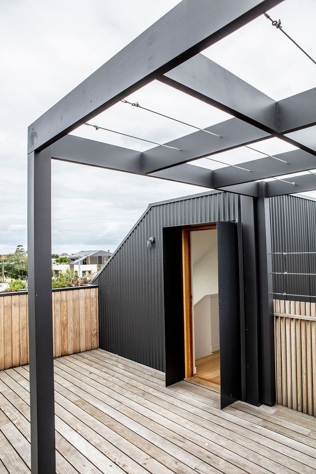 Exterior view of a contemporary rooftop or upper-level timber deck with a black steel pergola frame and tension cables overhead. The deck is enclosed by vertical timber privacy fencing and dark corrugated metal cladding, with an open doorway leading to an interior stair. Suburban rooftops and overcast sky are visible beyond.