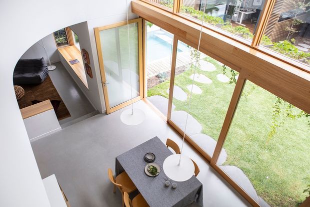 Elevated interior view of a contemporary dining area with a rectangular dining table and wooden chairs beside extensive floor-to-ceiling timber-framed glazing. The glazing overlooks a landscaped courtyard with lawn and stepping stones. A connected living area is visible through a large arched opening, and a mezzanine or upper-level edge with timber detailing appears above.