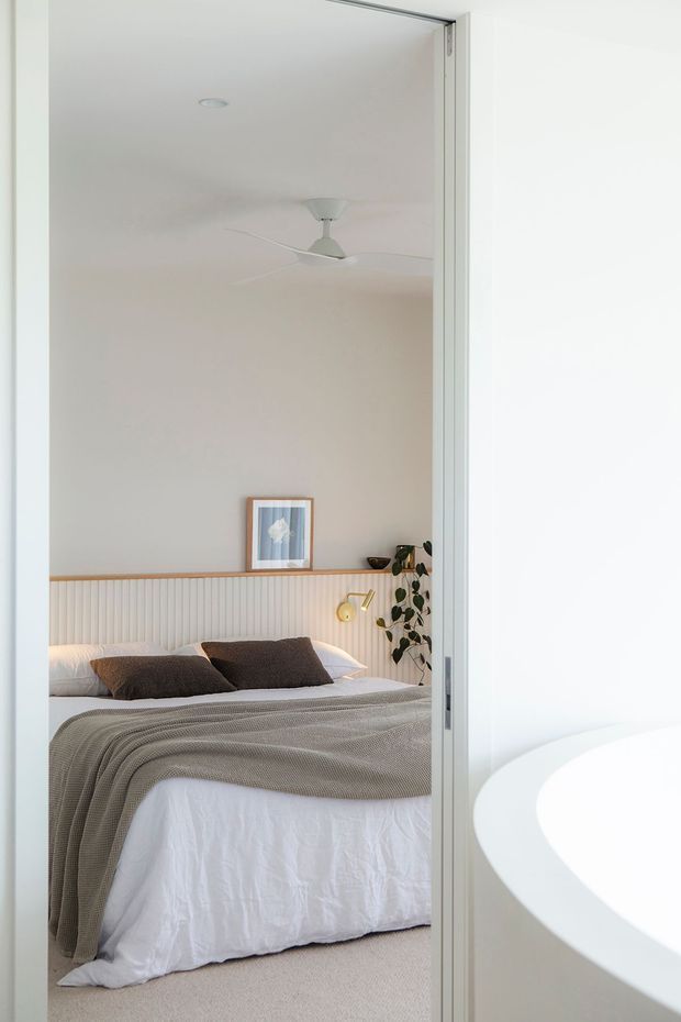 Bright, softly styled bedroom viewed through an open doorway, featuring a neatly made bed with white bedding and a taupe textured throw, dark neutral pillows, a vertical fluted wall panel behind the bed, pale warm walls, a ceiling fan, framed artwork, a small wall light, and a trailing indoor plant. The curved edge of a white freestanding bathtub is visible in the foreground at right, indicating an adjoining bathroom. Overall aesthetic is calm, minimal, and contemporary with warm neutral tones.