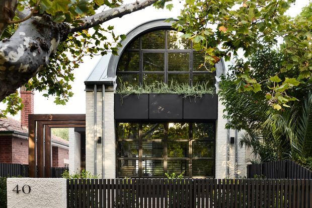 Front exterior view of a contemporary multi-level residence with a prominent arched, black-framed glazed facade, pale masonry walls, dark metal planter boxes, and a dark vertical-slat front fence. Mature leafy planting partially frames the building, with neighbouring brick residential structures visible at the sides.