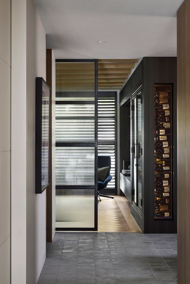 A sophisticated residential hallway or entry passage with dark built-in cabinetry, a tall integrated wine display, framed ribbed/frosted glass screens, and a glazed door leading toward a compact study or seating area. The passage combines grey textured floor tiles with warm timber flooring, exposed timber ceiling battens, dark joinery, and filtered natural light.