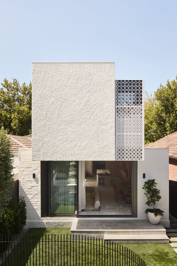 Front exterior view of a compact contemporary residence with a textured white rendered upper volume, patterned perforated privacy screen, large ground-floor glazing, and white-painted masonry walls. A small paved entry terrace, lawn, curved black metal fence, and mature planting frame the facade.