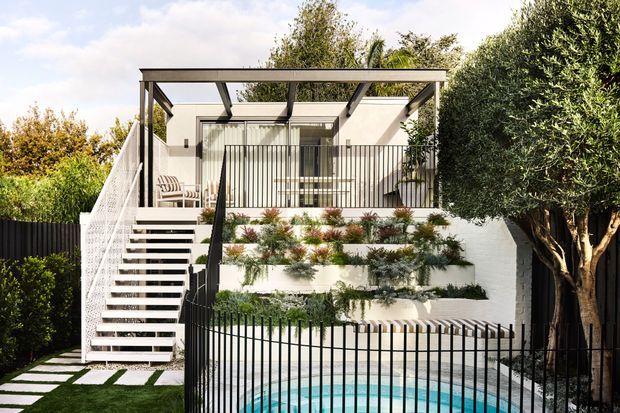 Exterior view of a contemporary multi-level home with a rooftop-style terrace and open pergola structure above the entry. A white rendered facade is paired with black metal balustrades, an external stair, tiered planted garden beds, and a partially visible swimming pool in the foreground. Mature greenery encloses the compact suburban outdoor space.