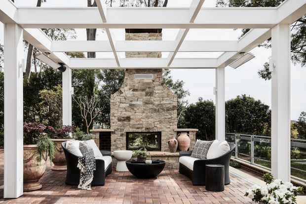 A refined outdoor terrace lounge beneath a white-painted pergola with a translucent or open grid roof. The space features black-and-white lounge chairs, a low black coffee table, a rustic stacked-stone fireplace, terracotta planters, brick paving, glass balustrades, and surrounding greenery. The composition suggests a sheltered outdoor entertaining area with an elevated, open outlook.