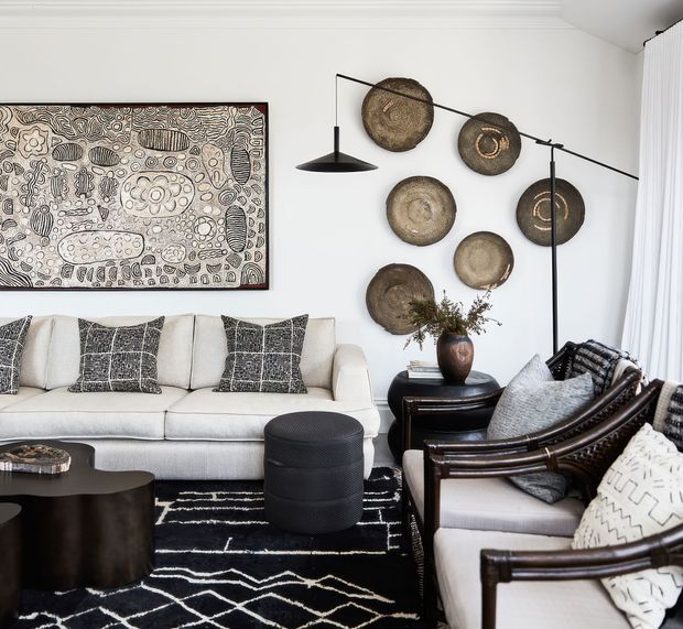 A refined living room with a white upholstered sofa, dark wood-framed armchairs, a black cylindrical ottoman, and a black-and-white geometric rug. The white walls feature a large monochrome artwork and a collection of circular woven wall pieces. A sculptural black floor lamp and side table with a vase add contrast, while sheer white curtains soften the room.