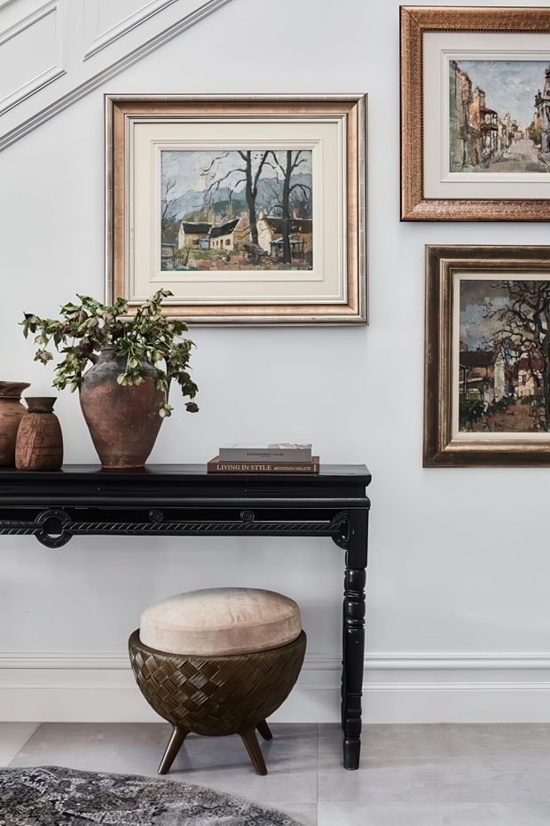 A refined interior vignette featuring a black console table with carved detailing against a pale grey paneled wall. Framed landscape artworks are arranged above the console, accompanied by a large rustic ceramic planter with trailing greenery, decorative pottery, and a small book. A woven dark wood or brown upholstered stool sits below, with pale tiled flooring and part of a patterned rug visible.