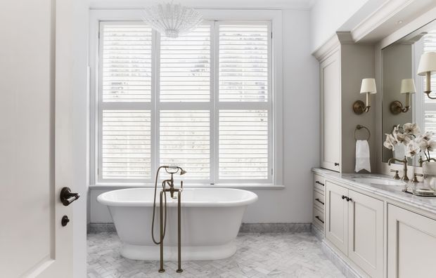 Bright, refined bathroom featuring a freestanding white bathtub beneath large plantation-shutter windows, a marble-look herringbone floor, and a built-in double vanity with pale cabinetry, dark hardware, marble countertop, wall sconces, and decorative mirrors. The room has a classic, elegant fit-out with abundant natural light.