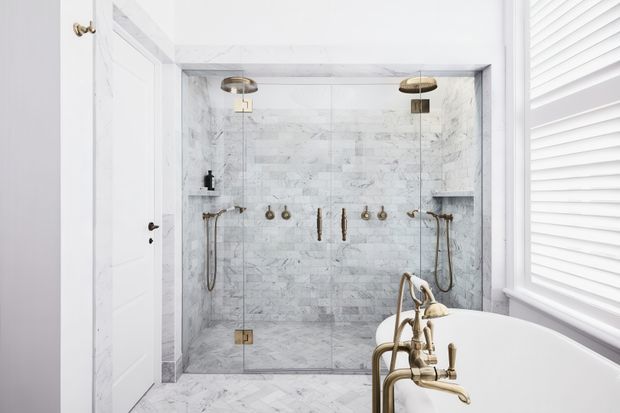 Bright, refined bathroom featuring a large walk-in glass shower lined in white and grey marble-look tile, dual shower fixtures, brass/gold hardware, and a freestanding white bathtub in the foreground. White paneled door and white plantation shutters reinforce the clean, classic look.
