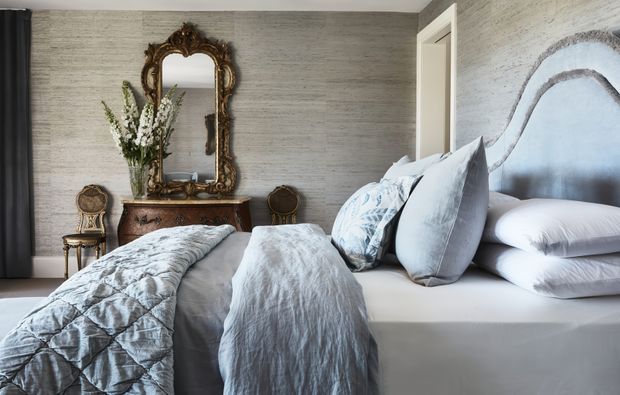 Elegant bedroom with twin beds dressed in pale blue-grey textured linens, set against a grey textured wall. An ornate antique-style gilt mirror hangs above a dark timber console, with classic accent chairs and a tall floral arrangement adding traditional character. A sculptural padded headboard and soft neutral palette create a refined, serene atmosphere.