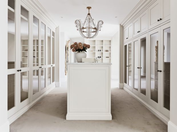 Bright, refined dressing room or walk-in wardrobe with extensive white built-in cabinetry, glass-front doors, mirrored panels, open shelving, and a central island. A sculptural chandelier hangs above the island, while neutral carpeting and white panelled joinery create an elegant, symmetrical interior.