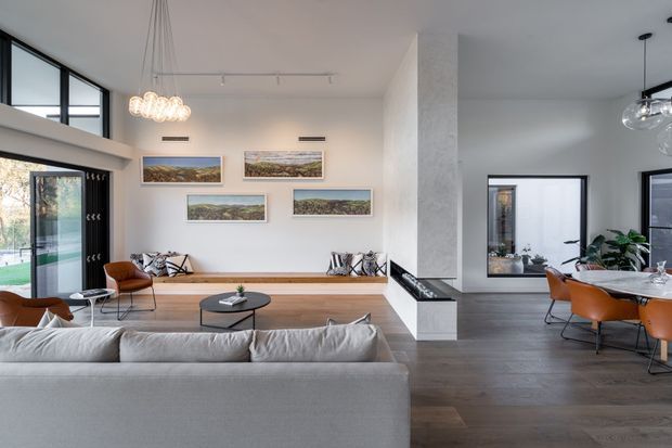 Bright open-plan contemporary living and dining area with a large pale sectional sofa in the foreground, timber flooring, a long built-in timber bench, sculptural fireplace surround, and expansive black-framed glazing. The adjacent dining area has a round table with orange upholstered chairs, while landscape artwork and indoor plants add visual interest. The room has a spacious, refined, neutral-toned appearance with high ceilings and abundant natural light.