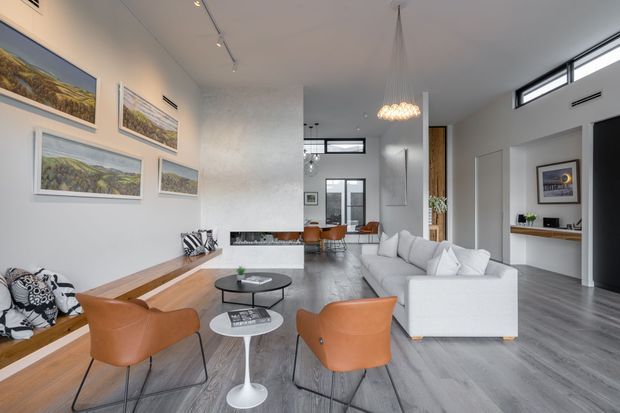 Bright, spacious contemporary open-plan living room with a white sectional sofa, leather lounge chairs, round coffee tables, wall-mounted artwork, and a linear timber bench along the left wall. The room features high ceilings, extensive glazing, polished grey flooring, recessed and track lighting, and a sculptural pendant above the seating area. A compact kitchen or bar area is visible at the rear/right.