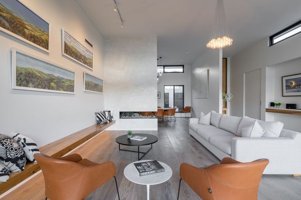 Bright, spacious contemporary living room with a white sectional sofa, cognac leather lounge chairs, a long built-in timber bench, round coffee tables, and a linear fireplace set into a textured feature wall. Track lighting and a sculptural pendant illuminate the room, while large framed landscape artworks and extensive glazing add visual interest. A dining area is visible beyond the living space.