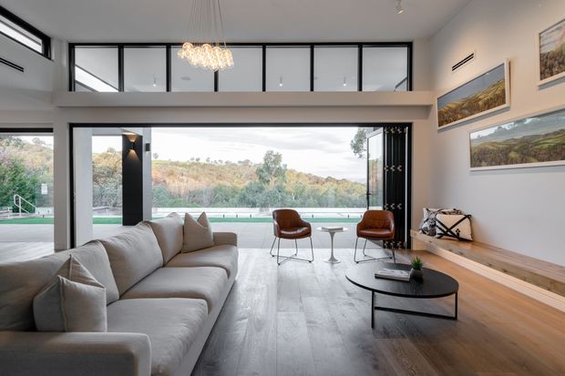 Contemporary open-plan living room with expansive floor-to-ceiling glazing and views toward a landscaped outdoor area. The room features a large light-grey sectional sofa, black oval coffee table, two brown leather lounge chairs, pale timber flooring, a long built-in timber bench, framed landscape artwork, and a sculptural pendant light. High glazing and a raised ceiling create a bright, spacious feel.