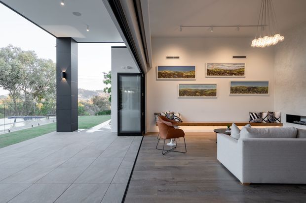 A refined contemporary living room opening directly onto a covered outdoor terrace through expansive black-framed glazing. The interior features pale timber flooring, a white sectional sofa, a compact timber dining table with leather chairs, built-in bench seating, framed landscape artwork, recessed ventilation, and a sculptural suspended light. The terrace uses large-format grey paving and overlooks a landscaped lawn and pool area.