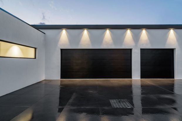 Exterior view of a sleek contemporary garage facade with two dark sectional garage doors, white rendered walls, linear roofline, and evenly spaced wall-mounted downlights. The glossy dark paved driveway reflects the lighting, creating a dramatic evening atmosphere.
