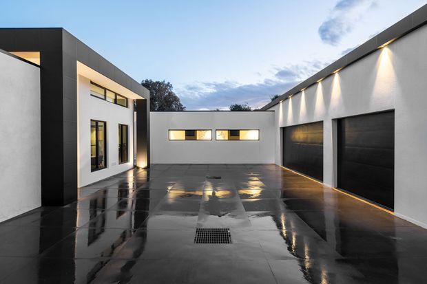 A contemporary courtyard-style residential exterior viewed from a paved driveway or forecourt. White rendered walls are framed with bold black cladding and trim, with narrow horizontal windows, warm exterior lighting, and two dark garage doors. The glossy dark paving reflects the building and lights, creating a dramatic evening atmosphere.