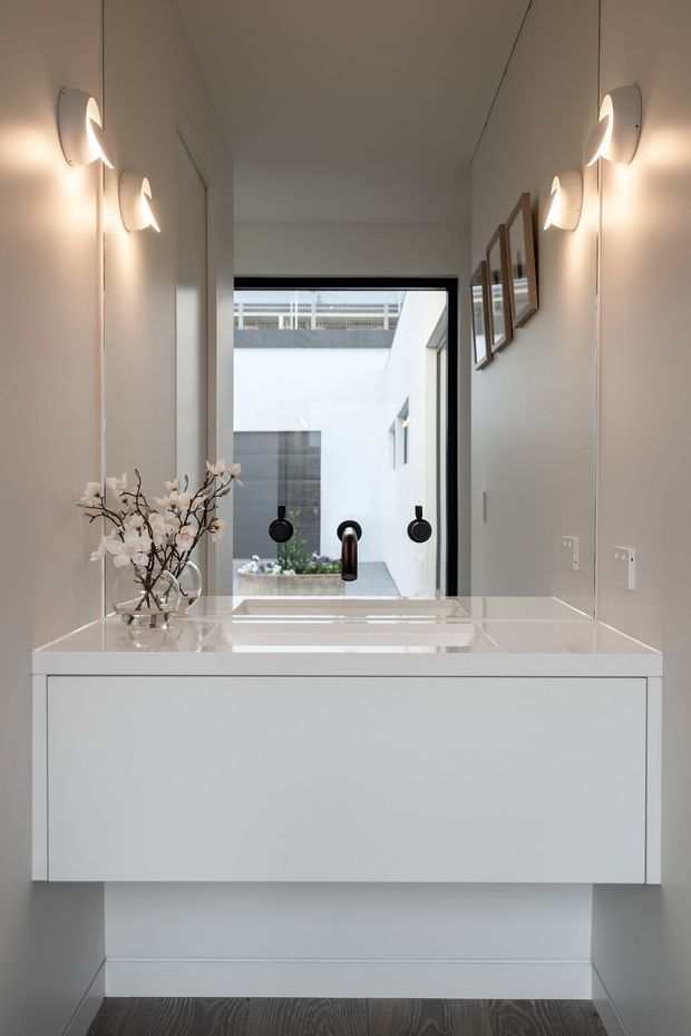 A refined minimalist hallway or entryway with a floating white built-in console and integrated washbasin beneath a large mirror. The space features pale grey walls, contemporary sculptural wall sconces, framed artwork, dark timber flooring, and a view toward a bright internal courtyard through glazing. Black plumbing fixtures provide contrast against the white vanity and light interior.