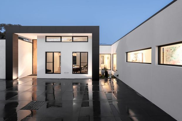 Contemporary single-level courtyard house viewed from an outdoor paved courtyard at dusk. The composition features crisp white rendered walls, dark charcoal framing and flat roof forms, black-framed windows, warm timber cladding at the entry, and glossy dark paving reflecting the building and evening light. Interior artwork and furnishings are visible through the glazing.