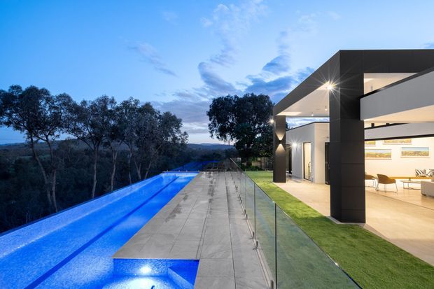 Contemporary luxury house exterior at dusk, featuring a long illuminated swimming pool beside a broad paved terrace with frameless glass balustrading. The building has strong rectilinear volumes, dark cladding, extensive open glazing, and warm-lit interior spaces, set among mature trees with distant landscape views.