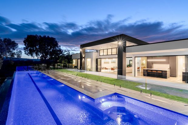 Twilight exterior view of a contemporary single-storey residence with a dramatic low-pitched roof and extensive glazed openings. A long illuminated swimming pool and paved terrace lead toward the open-plan interior, where a kitchen and living area are visible. The property features clean geometric forms, dark structural accents, pale walls, and landscaped lawn.