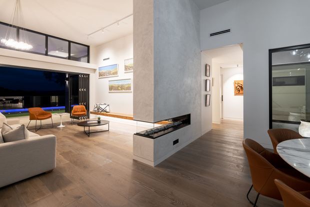 Open-plan contemporary home interior featuring a living room, dining area, and hallway arranged around a central double-sided fireplace. The space has warm timber flooring, white walls, large dark-framed glazing, sculptural pendant lighting, a neutral sectional sofa, orange accent chairs, and a marble dining table. A dark outdoor view is visible through the living-room glazing, while framed artwork and a glazed room opening add visual detail.