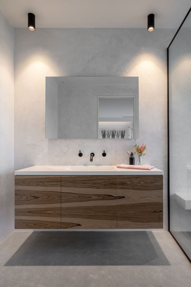 A contemporary bathroom vanity with a large floating timber-grain cabinet, white integrated benchtop and basin, black wall-mounted tapware, and a frameless mirror. The room has textured light grey plaster walls, a grey concrete-look floor, black cylindrical ceiling spotlights, and a large glass shower enclosure on the right. The palette is restrained and modern, with warm timber cabinetry providing contrast.